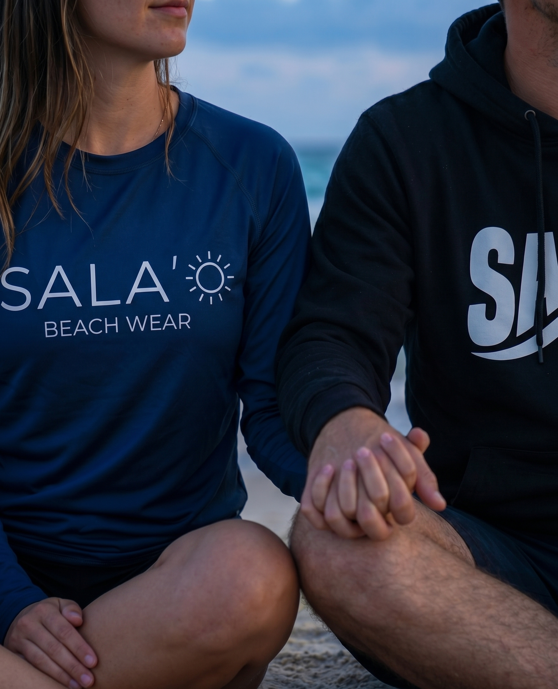 Two people sitting together wearing Sala'o Beach Wear clothing with a scenic background.